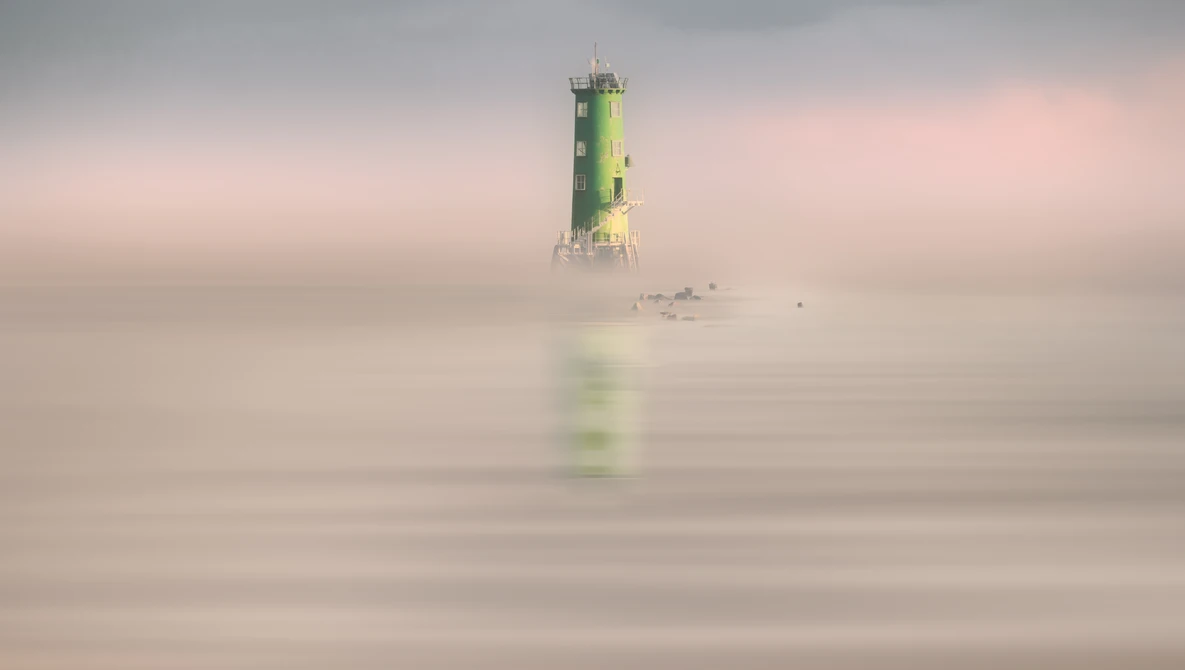 Green and yellow lighthouse emerging from thick fog over calm water.