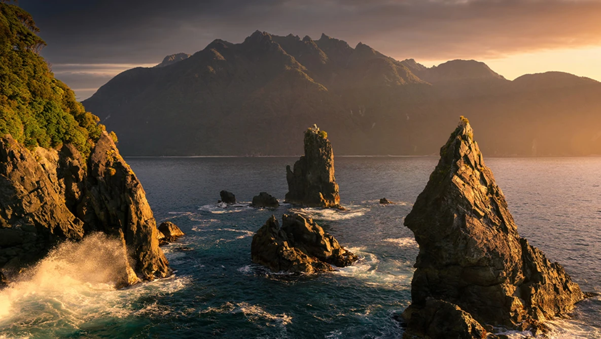 Dramatic rocky sea stacks catching golden sunrise light with misty mountains across the water.
