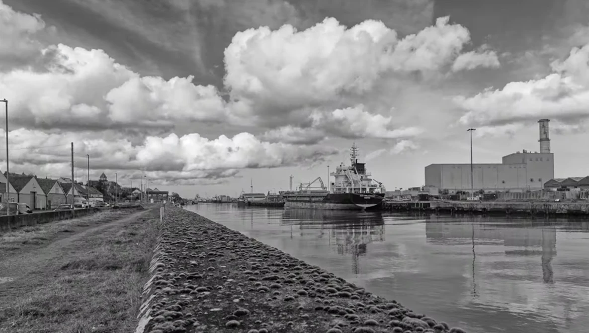 Industrial cargo ship moored alongside a stone quay with residential buildings and modern structures in the background under dramatic cloudy sky.