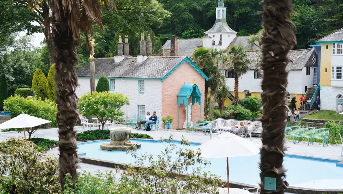 Coastal village with pastel buildings, palm trees framing the composition, and a swimming pool in the foreground.