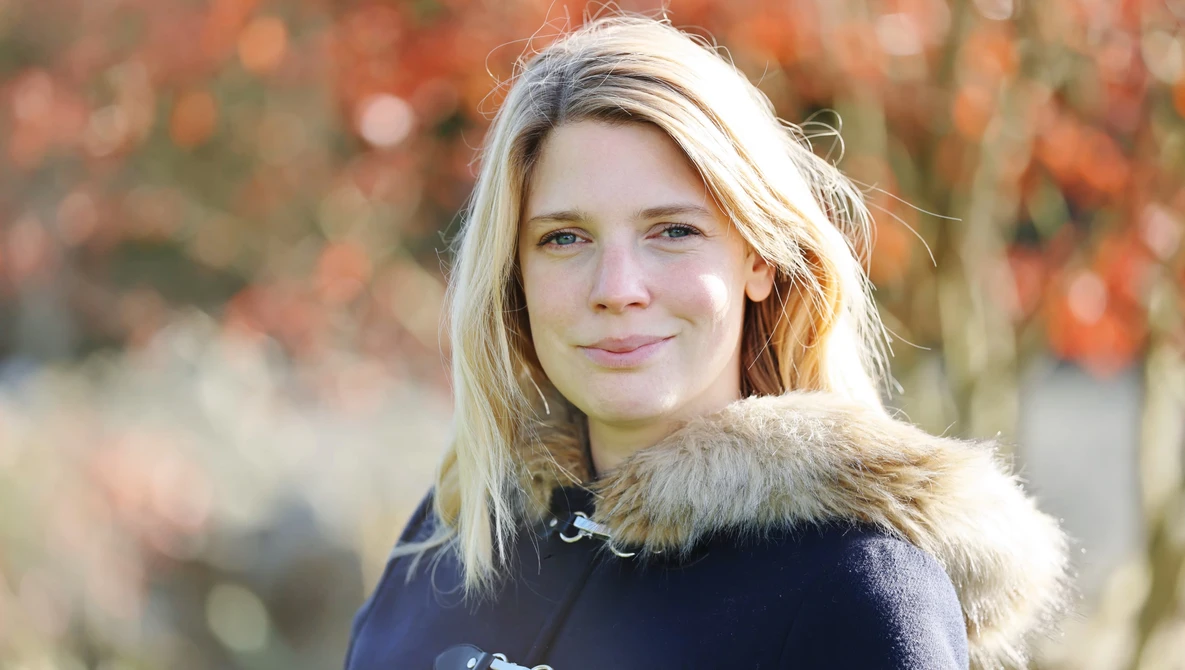 Young woman with blonde hair wearing a dark sweater with fur collar, photographed outdoors with autumn foliage in soft focus behind her.