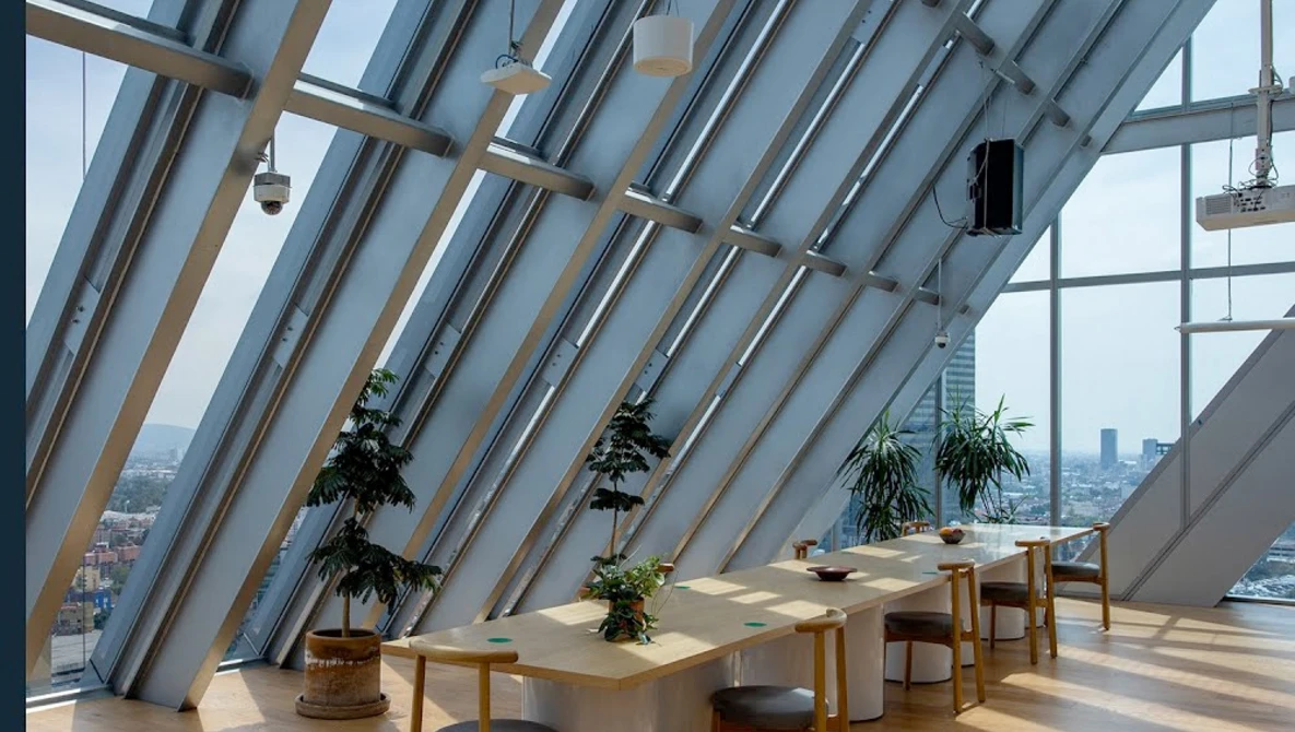 Modern interior with angled glass ceiling, wooden structural beams, suspended plants, and minimalist furniture overlooking a city skyline.