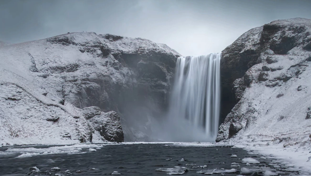 Tall waterfall cascading between snow-covered rocky cliffs into a dark pool of water.