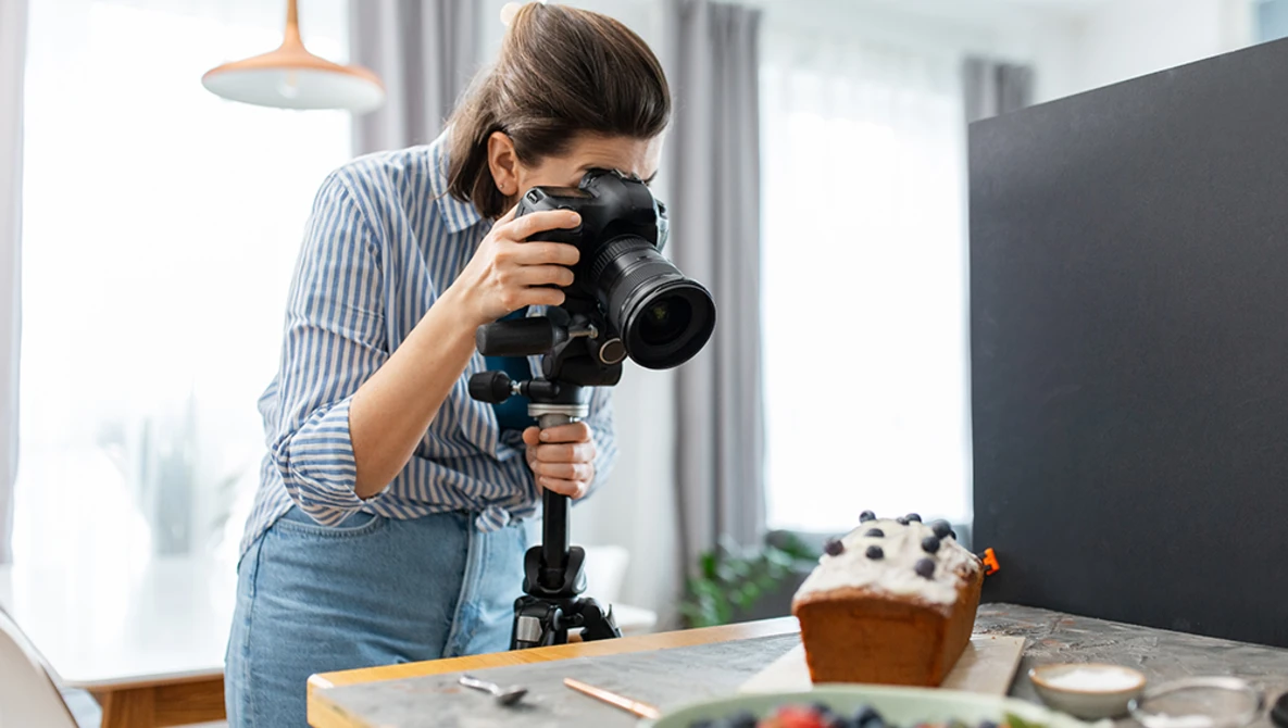 Female videographer operating a camera on a stabilizer rig while filming a styled food scene indoors.