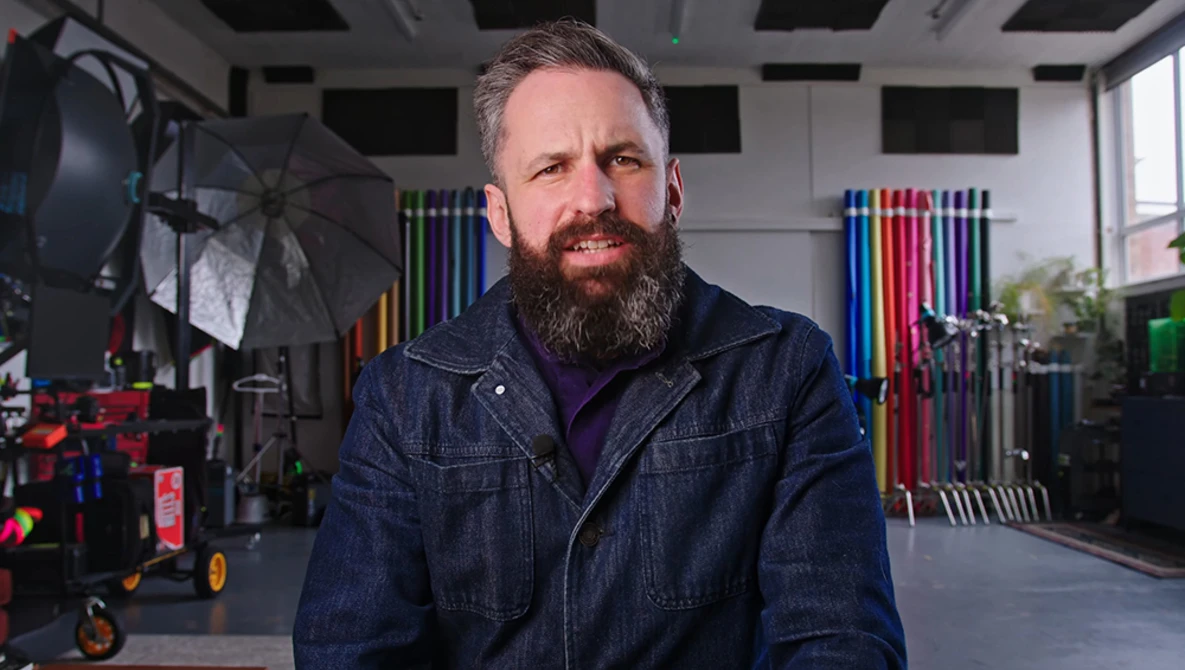 Bearded man in dark denim jacket standing in a professional studio with lighting equipment and colored gels visible in the background.