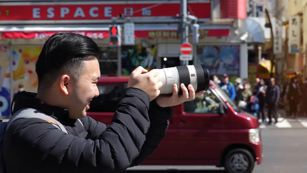 Photographer holding a telephoto lens up to his face while shooting on a busy urban street.