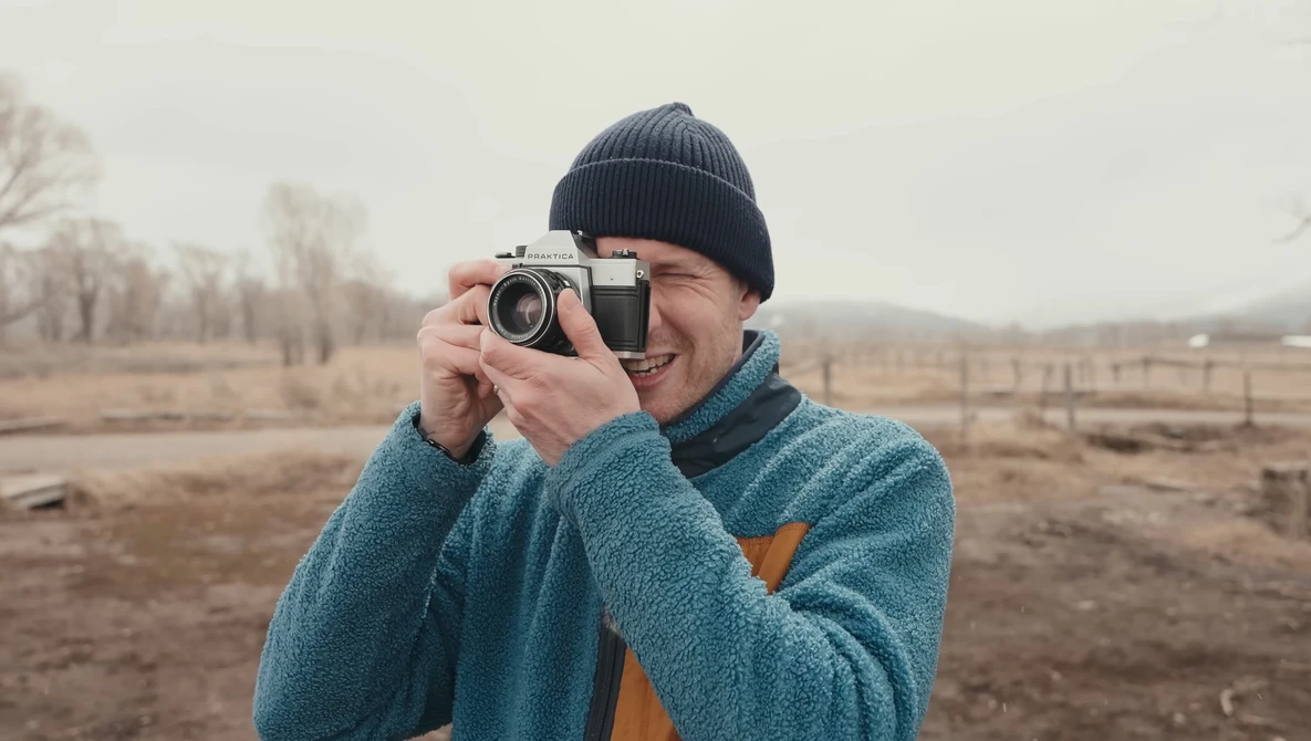 Smiling photographer holding a vintage rangefinder camera at eye level in an open field.