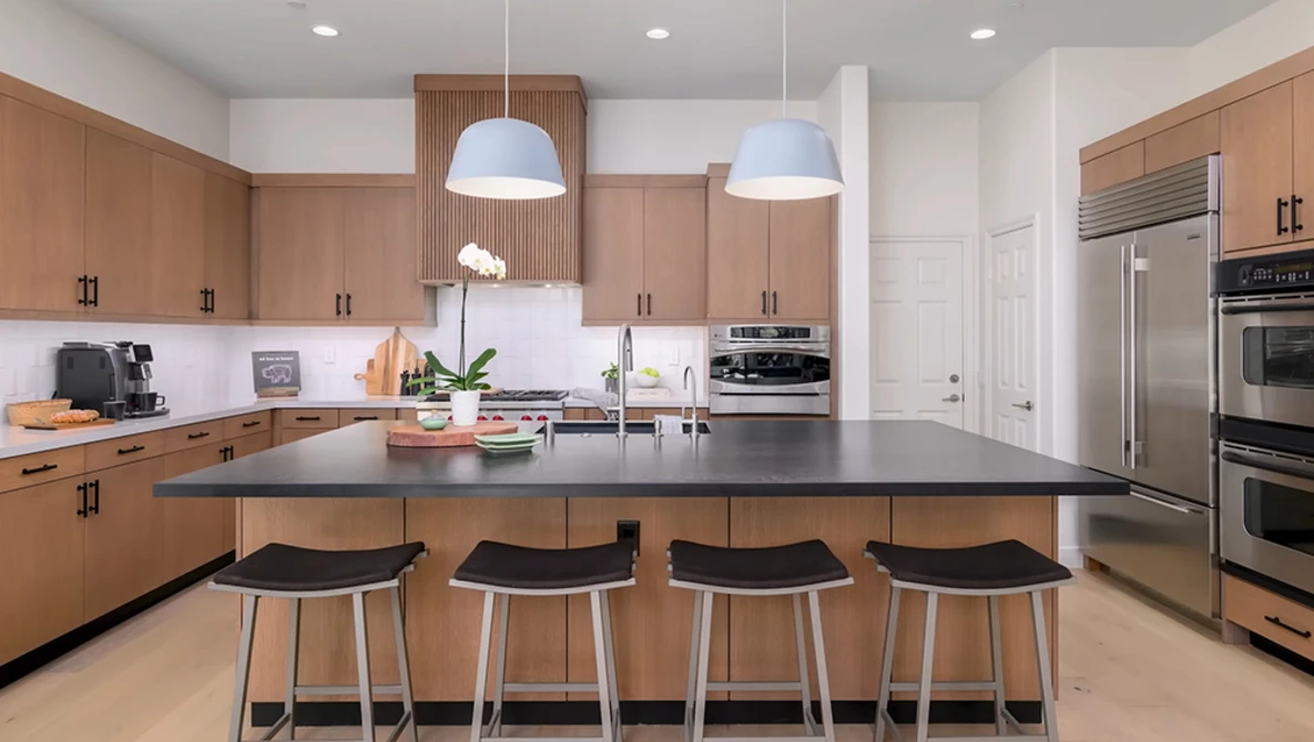 Modern kitchen with wood cabinetry, black island countertop, and four black bar stools under pendant lighting.