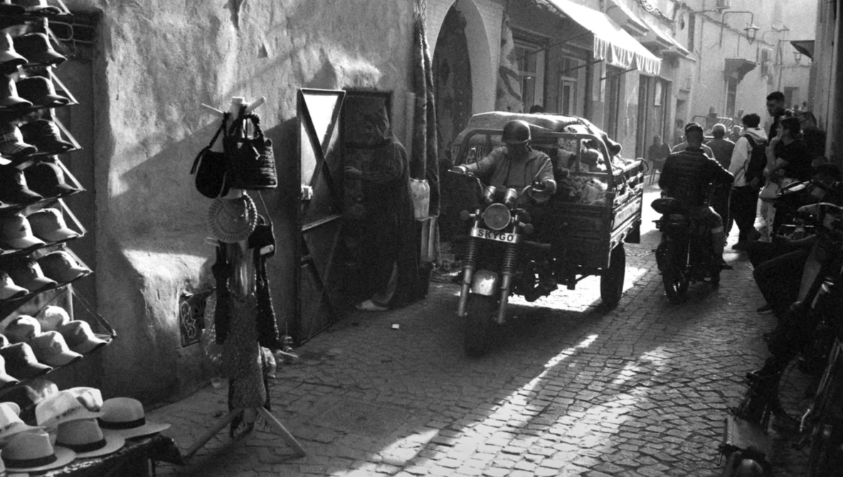 Black and white street scene with vintage car parked among pedestrians in narrow cobblestone alley.