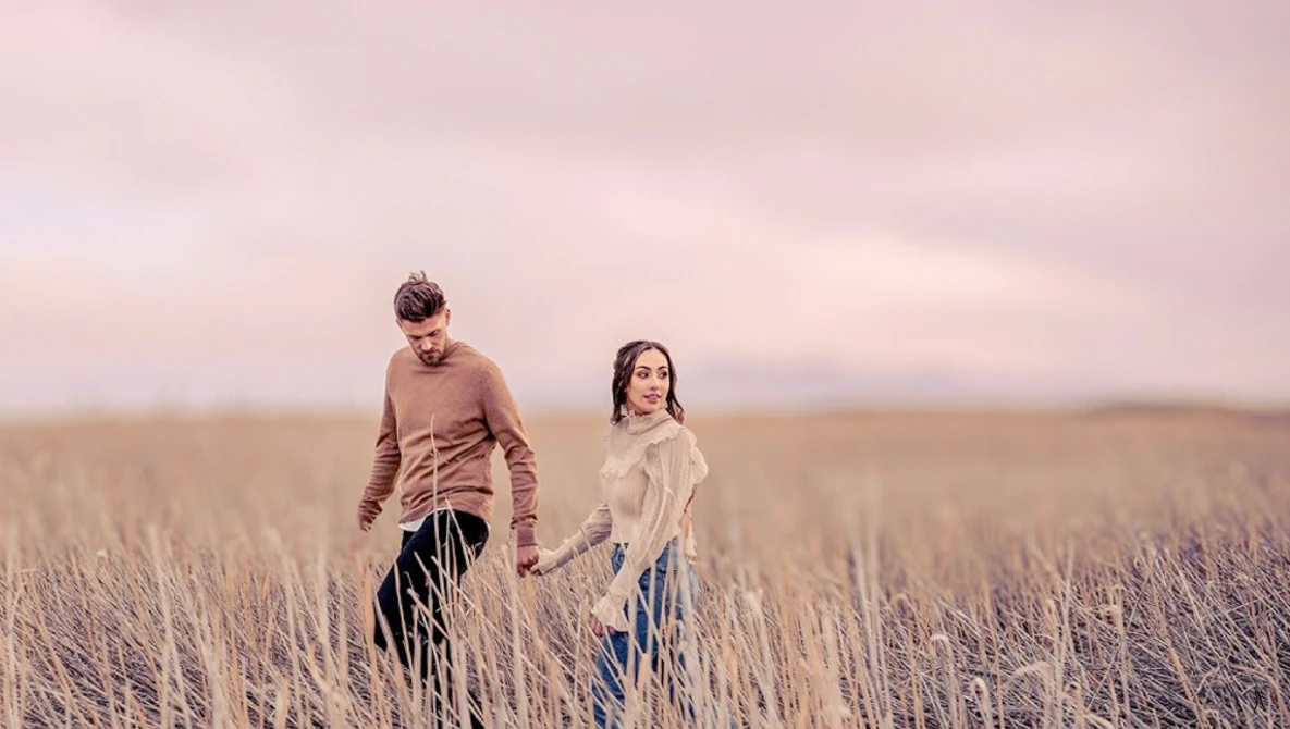 Two people walking through a dried grass field at dusk under a pink-toned sky.