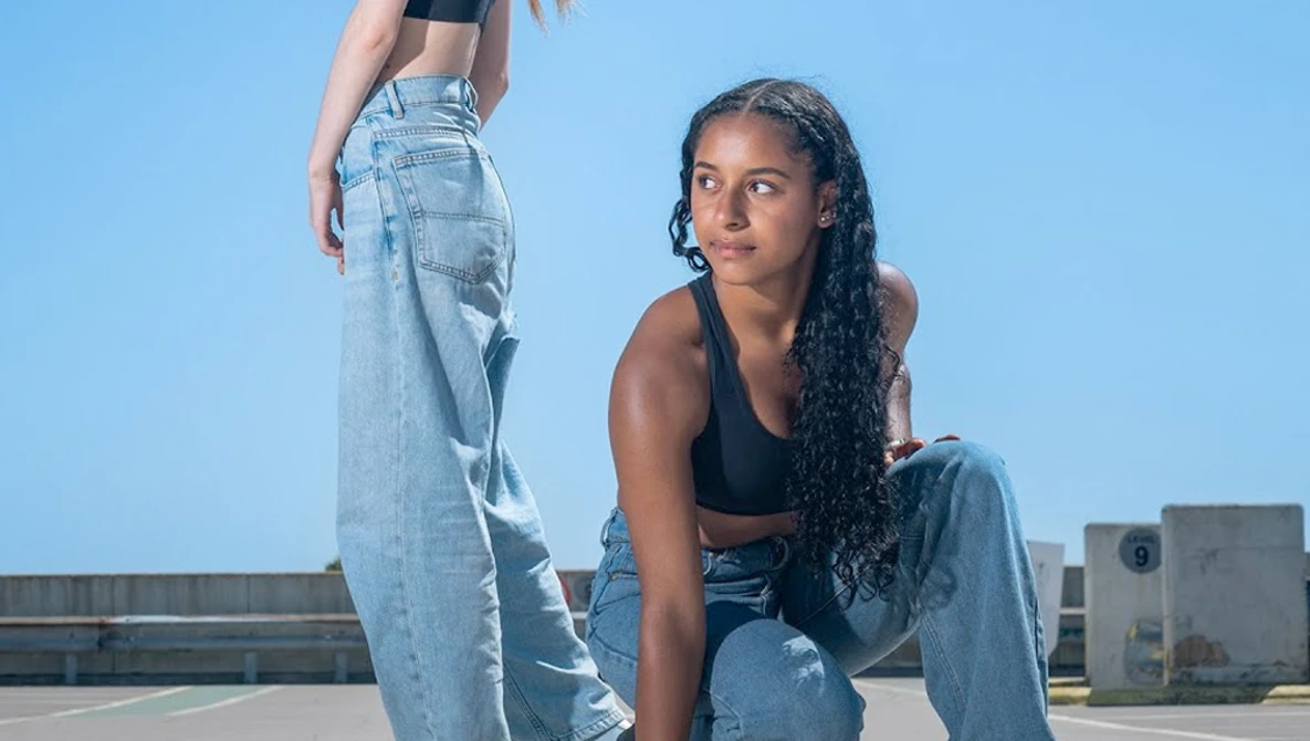 Two women posing on a rooftop against a clear blue sky, wearing distressed denim jeans and crop tops.
