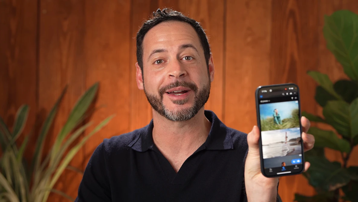 Man in black polo shirt holding smartphone displaying photo app interface against wooden wall with potted plants.