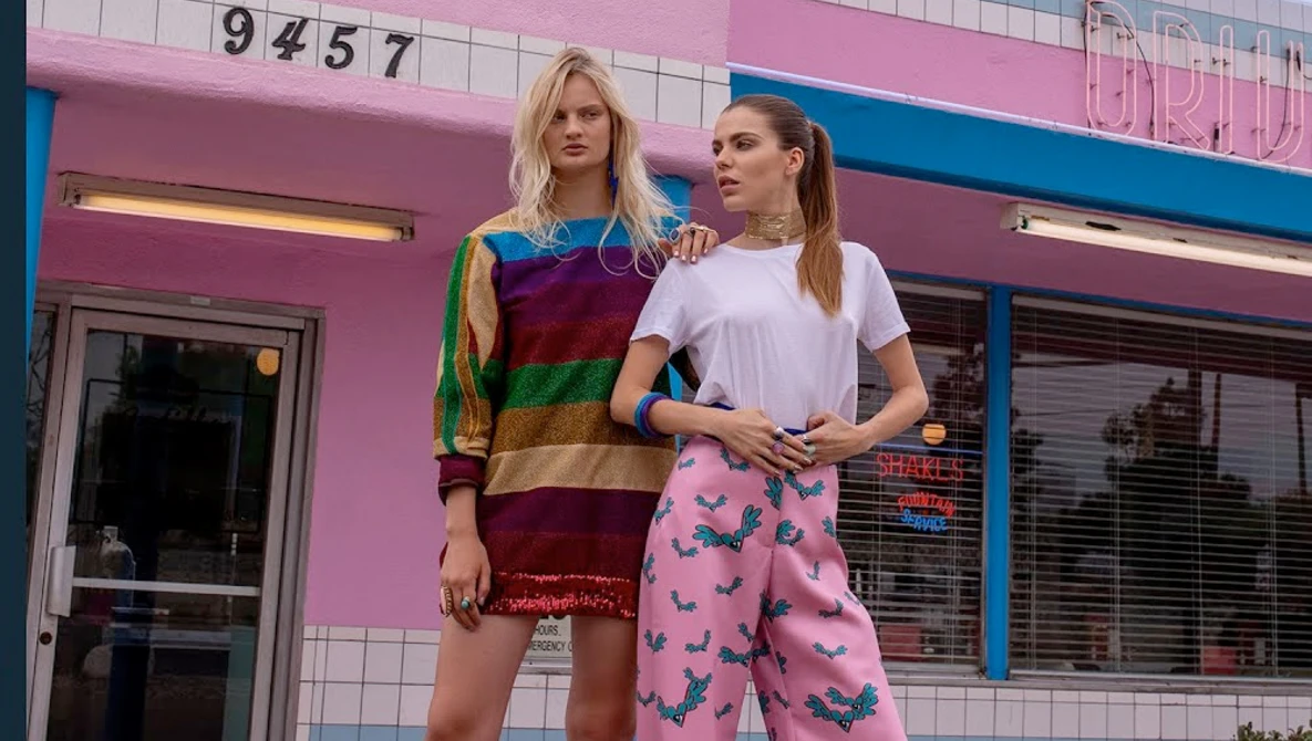Two women posing against a bright pink storefront with blue trim and white tile details.