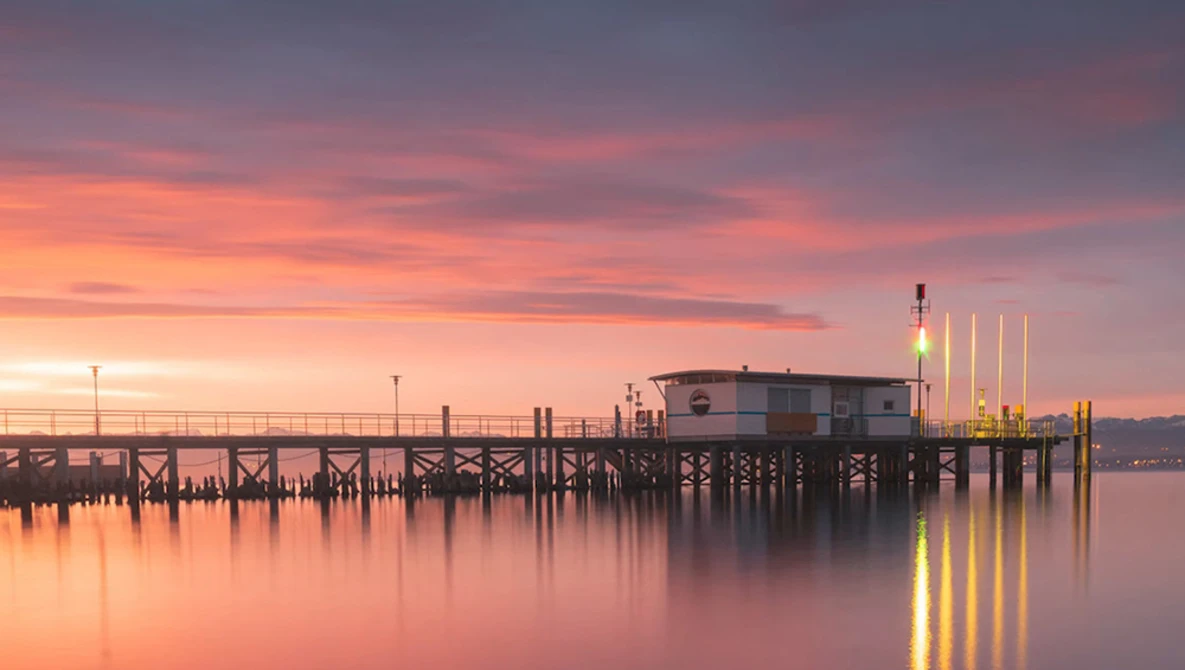 Wooden pier extending into still water at sunset with warm pink and orange sky reflecting on the surface.