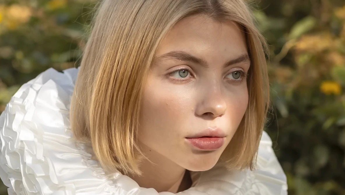 Young woman with shoulder-length blonde hair wearing a white textured garment, photographed outdoors with soft natural lighting.