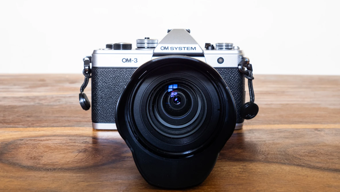 OM System OM-3 rangefinder camera with attached zoom lens on wooden surface.