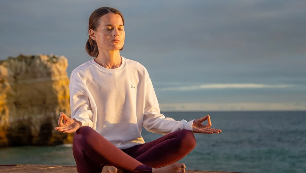 Woman in meditation pose overlooking ocean with rock formations in background.