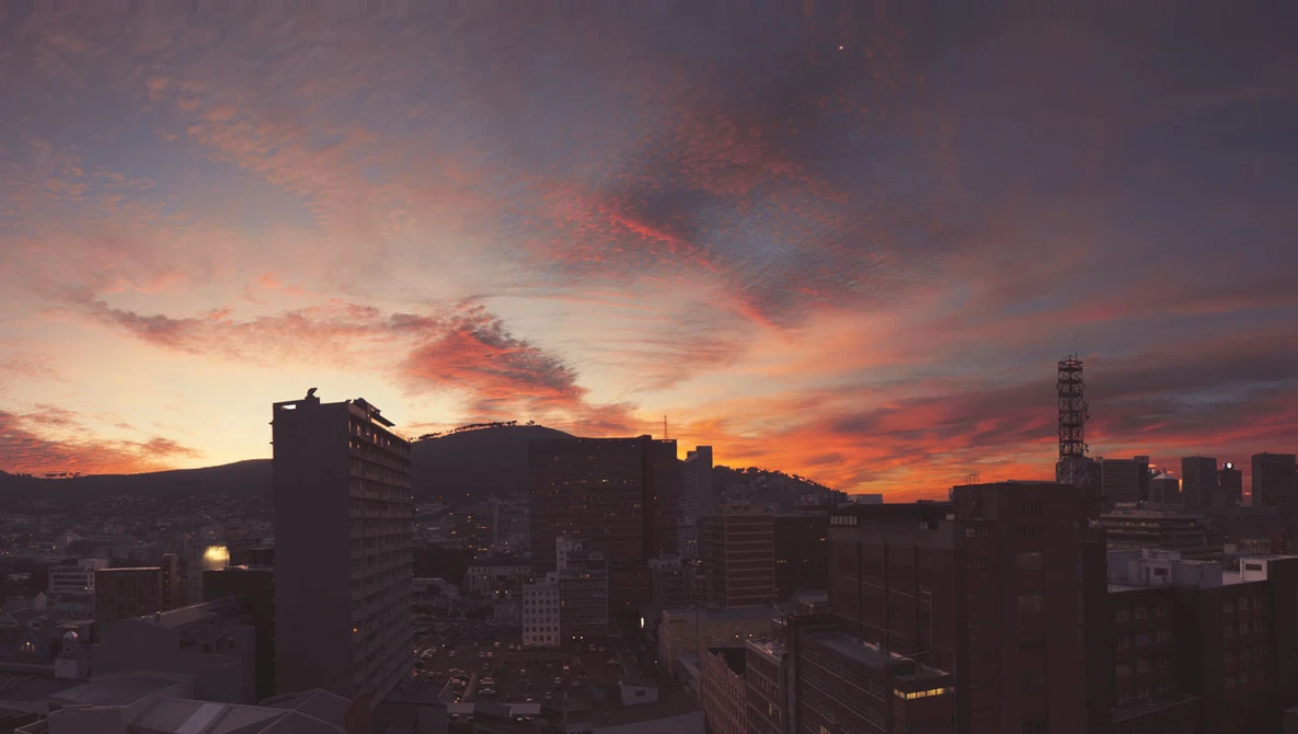 Urban skyline at sunset with warm orange and purple tones lighting silhouetted buildings and a distinctive peaked mountain.