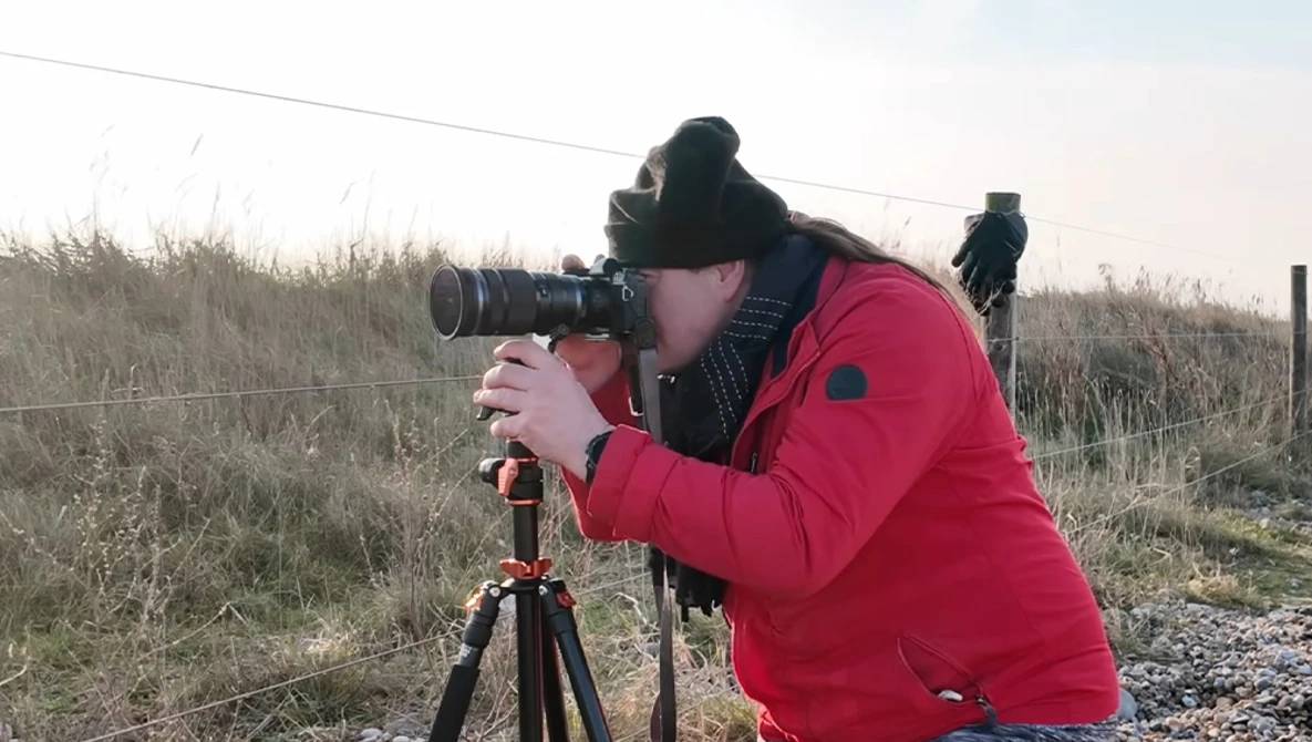 Photographer in red jacket looking through telephoto lens mounted on tripod in rural landscape.