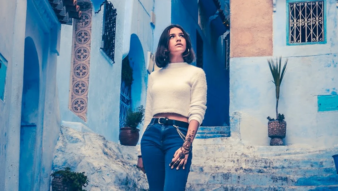 Woman in cream sweater and dark jeans standing against weathered blue and white building wall.