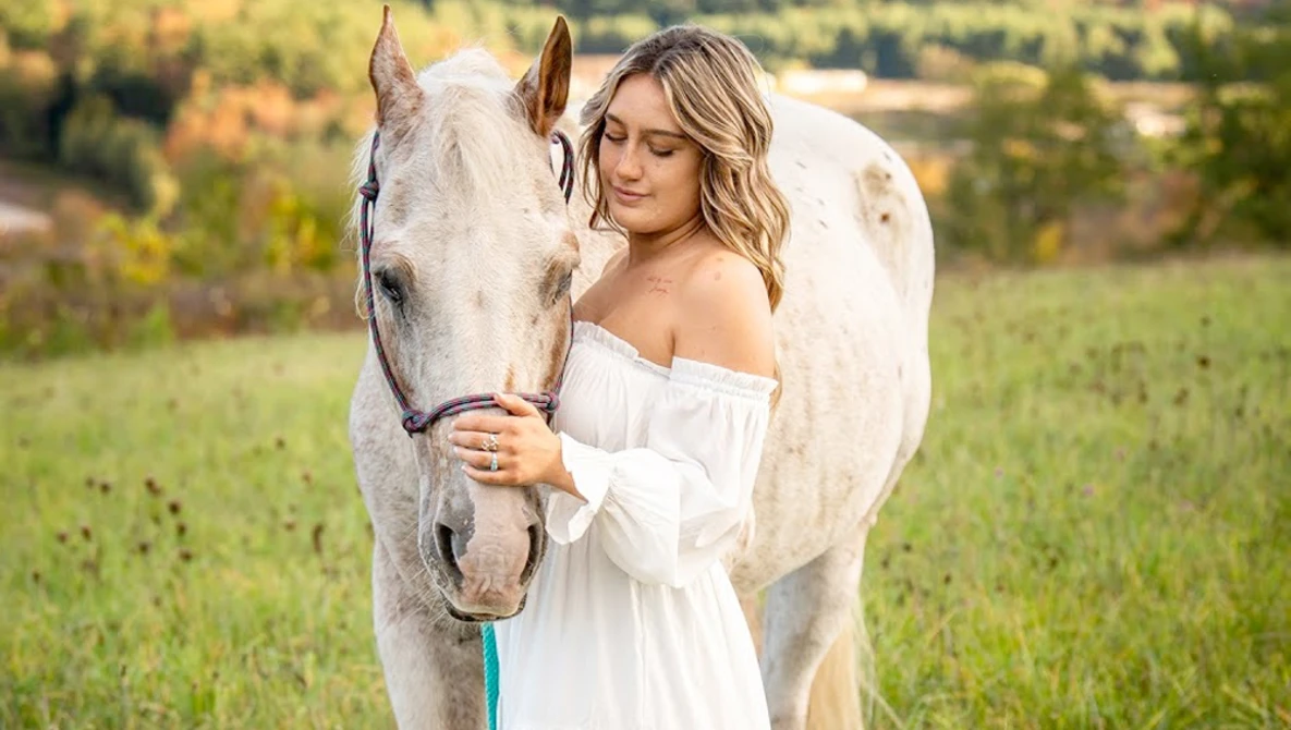 Woman in off-shoulder white dress standing beside a gray horse in a pastoral field.