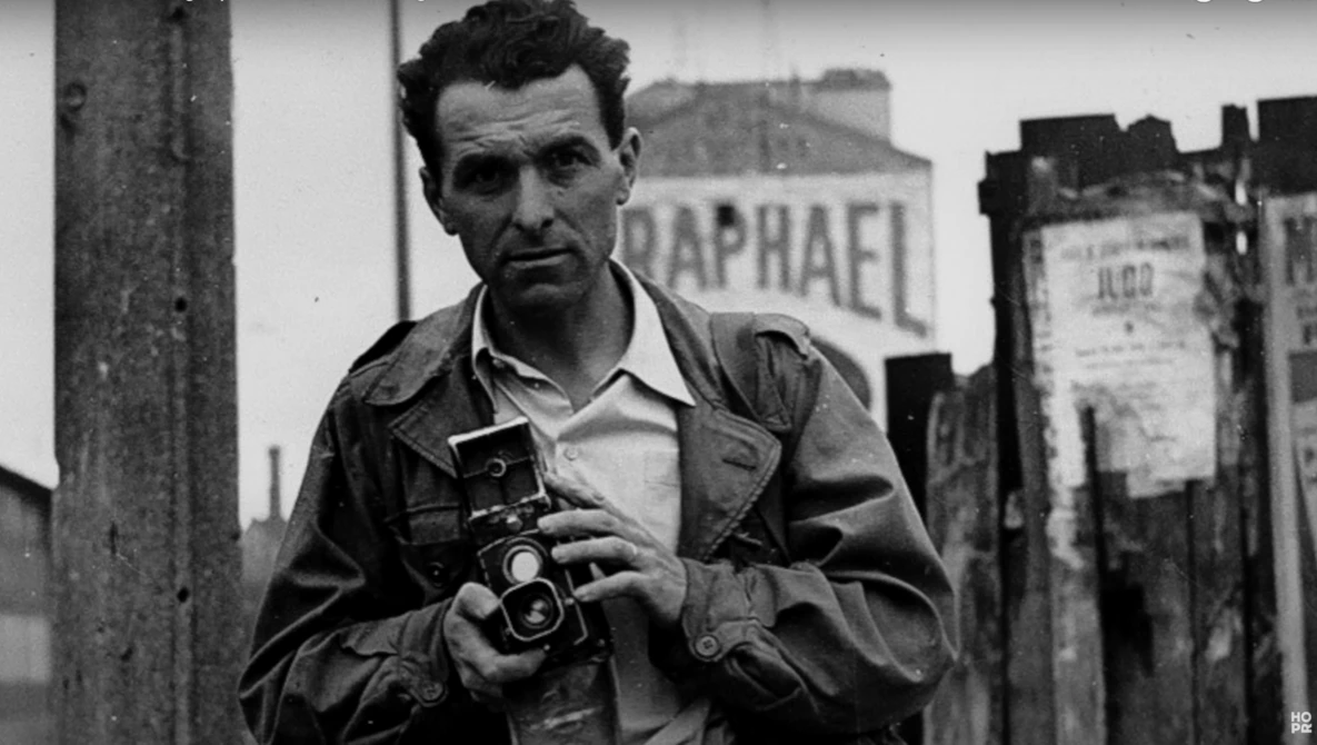 Man holding a vintage rangefinder camera in an urban setting with weathered buildings behind him.