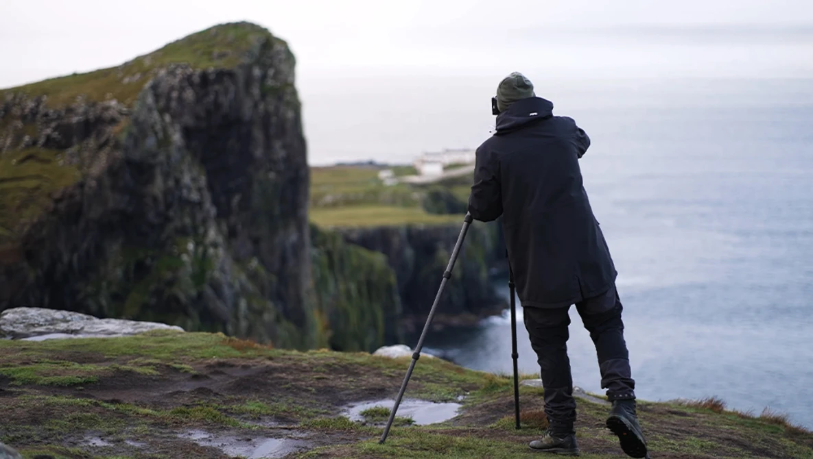 Hiker with trekking poles standing on mossy cliff edge overlooking ocean and coastal landscape.