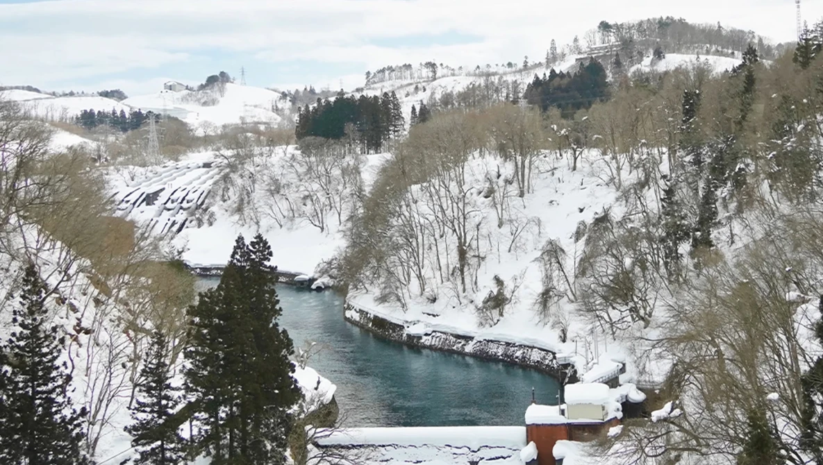 Snow-covered mountain landscape with a turquoise river flowing through a valley lined with evergreen and bare deciduous trees.