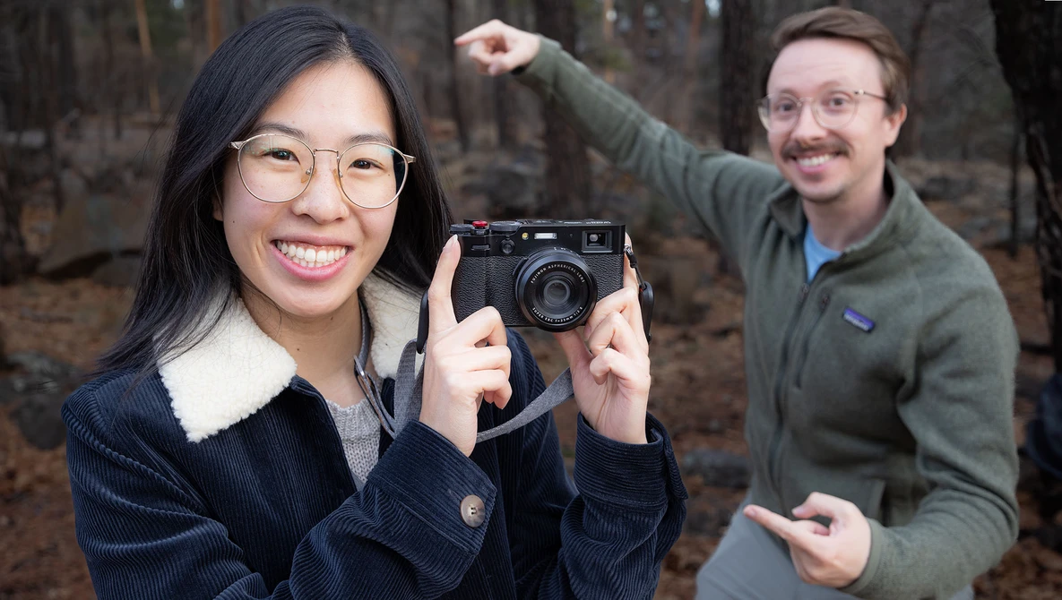 Woman holding a rangefinder camera while smiling at the camera, with a man gesturing enthusiastically in the background outdoors.