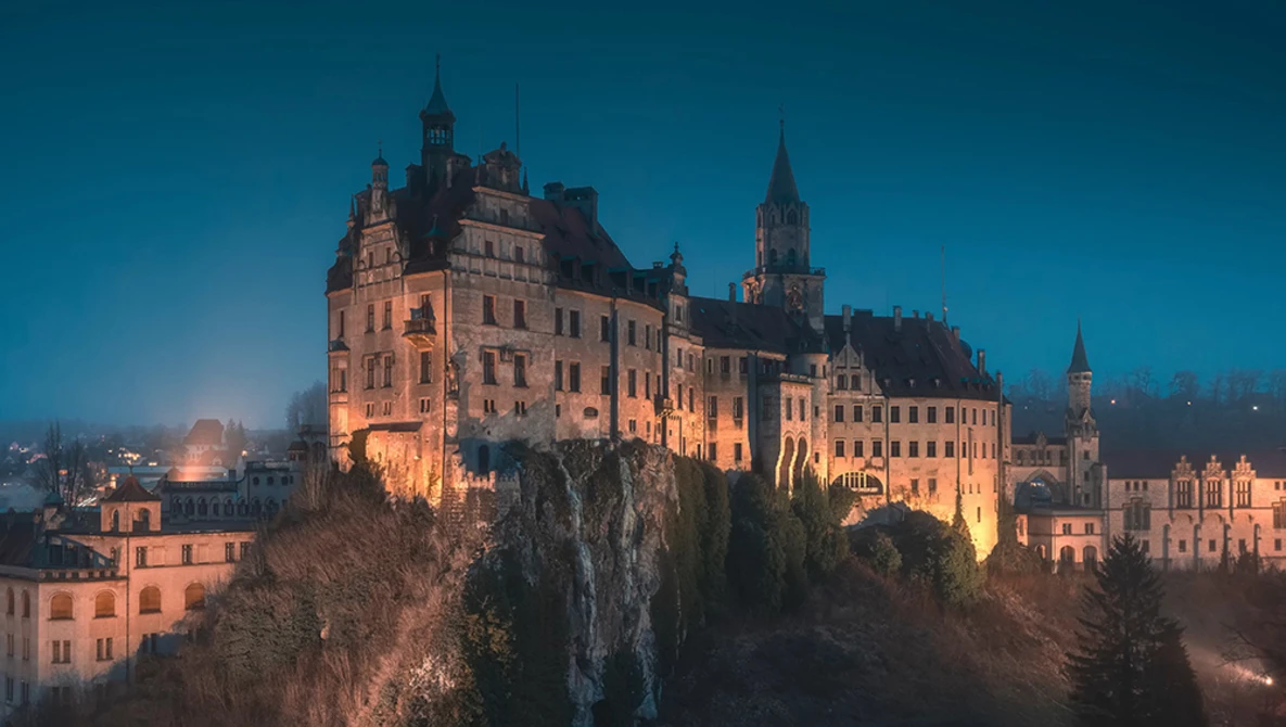 Illuminated castle perched on a forested cliff at twilight under a deep blue sky.