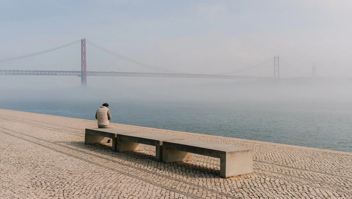 Solitary figure sitting on a bench facing a suspension bridge shrouded in fog.