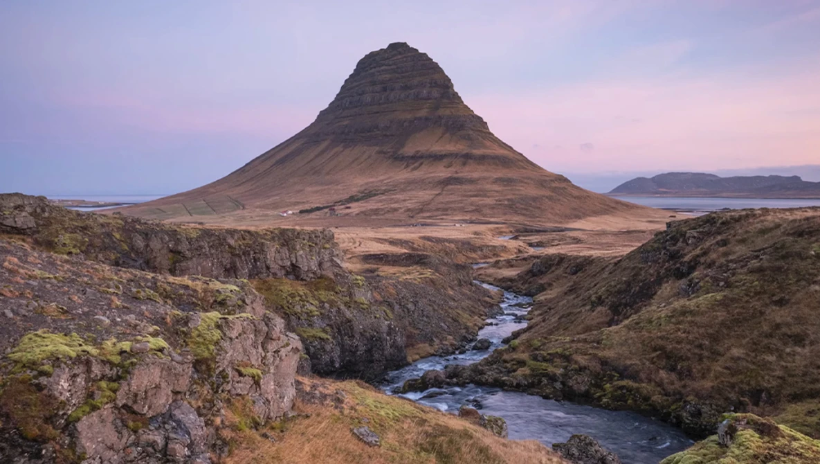 Kirkjufell mountain with snow-dusted slopes rising above a braided river valley at dusk.