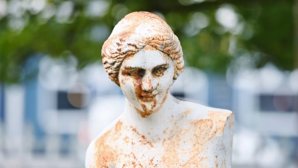 Weathered stone bust sculpture with patina and erosion marks, photographed outdoors against blurred background.