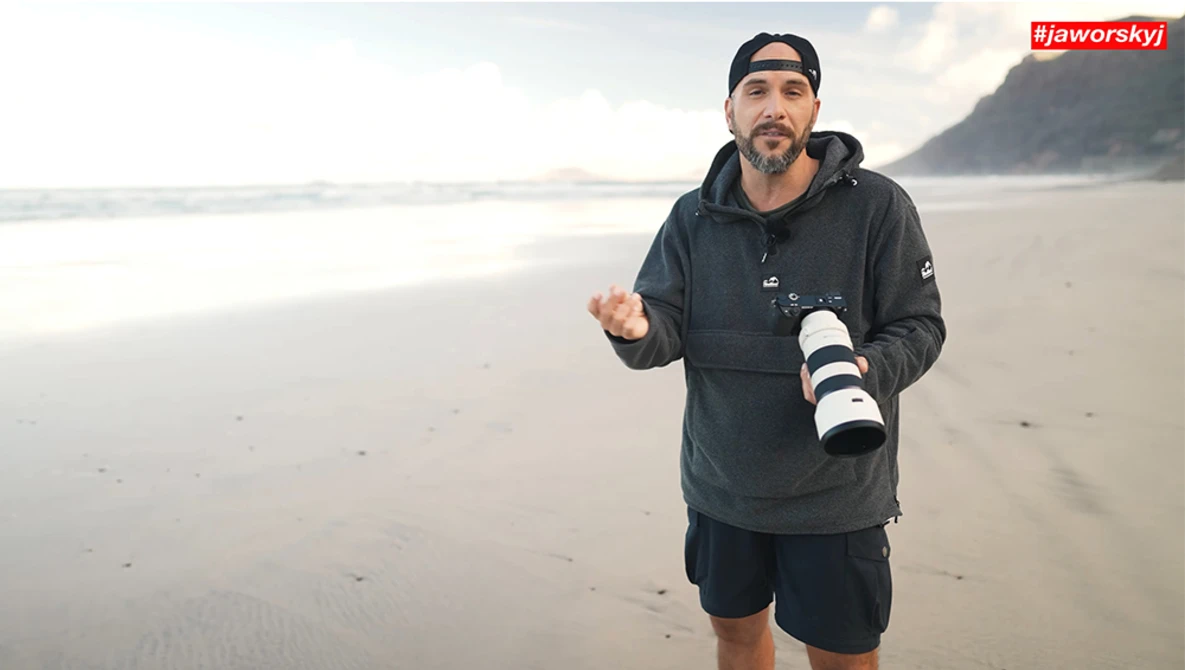 Photographer holding a telephoto lens on a misty beach while gesturing during discussion.