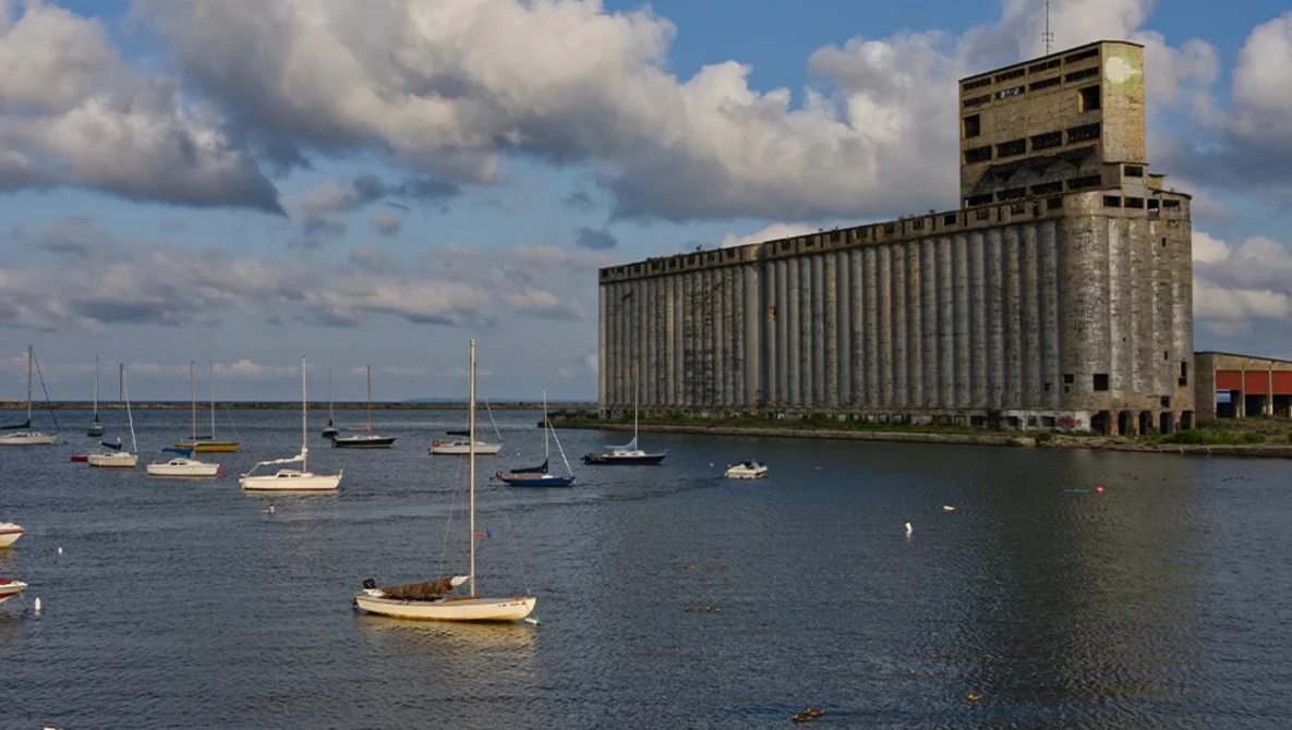 Sailboats moored in calm blue water near a large industrial grain elevator building under partly cloudy skies.