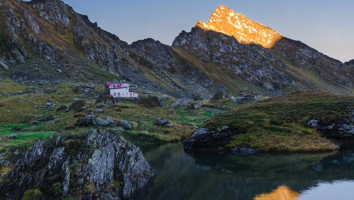 Alpine valley landscape with a pink building nestled between rocky peaks and a reflective pond in the foreground.