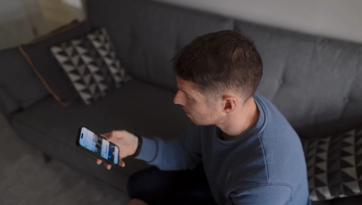 Man seated on a gray sofa looking at a smartphone screen.