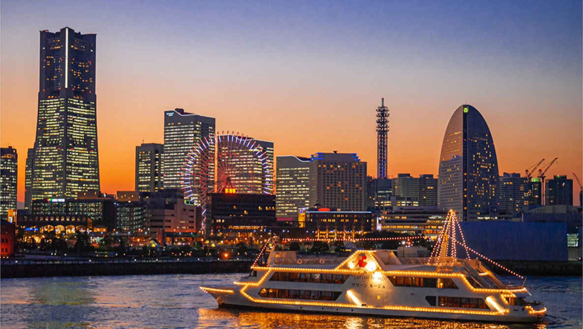 Illuminated ferry boat on water at dusk with city skyline and glowing buildings in background.