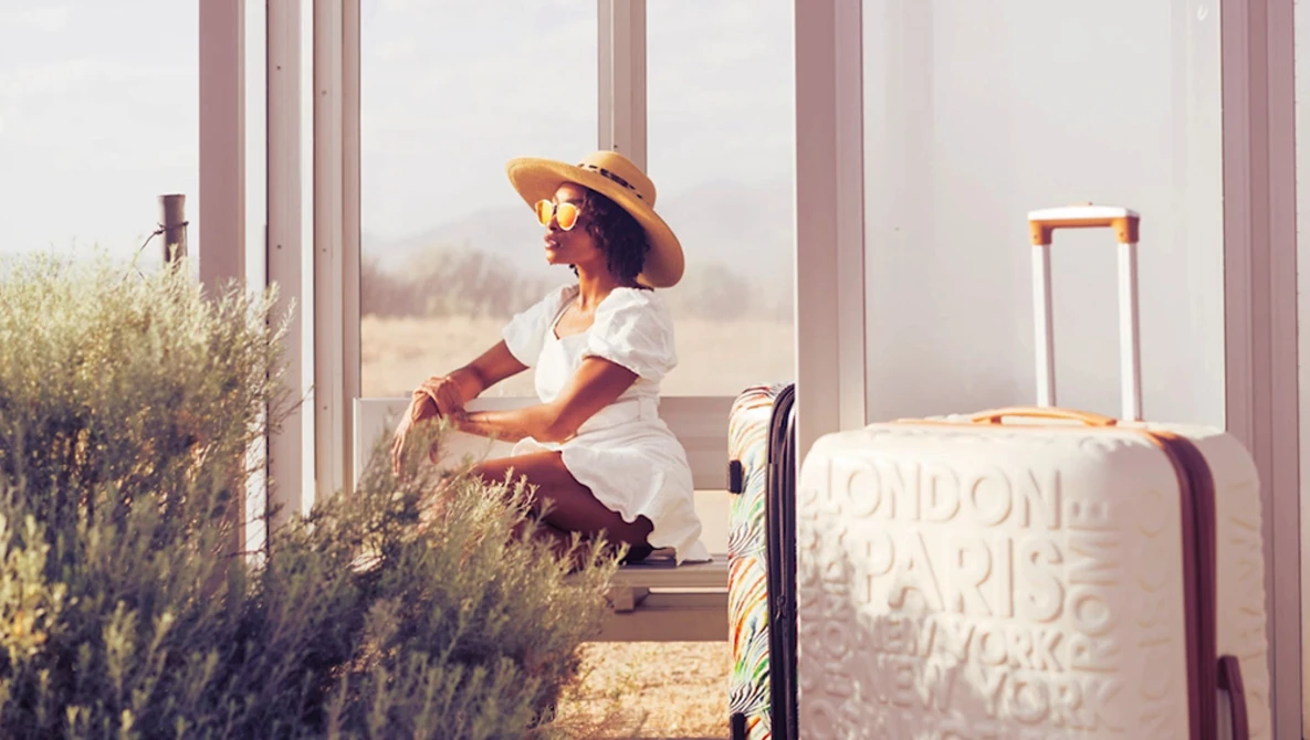 Woman in white dress and straw hat sitting by large windows with lavender plants and luggage beside her.