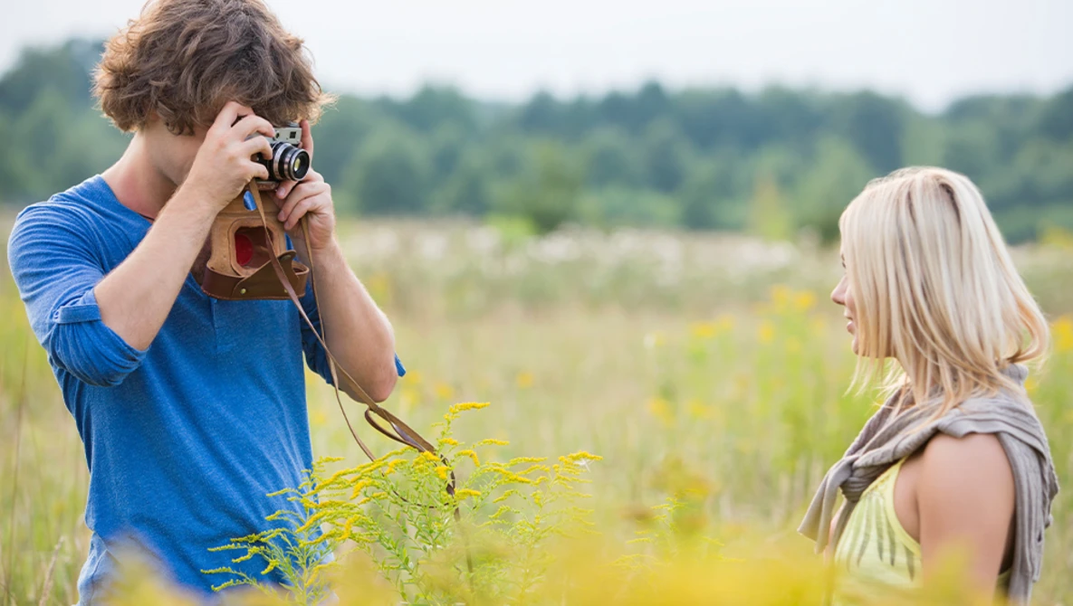 Young man in blue shirt photographing a woman in a field with a vintage camera.