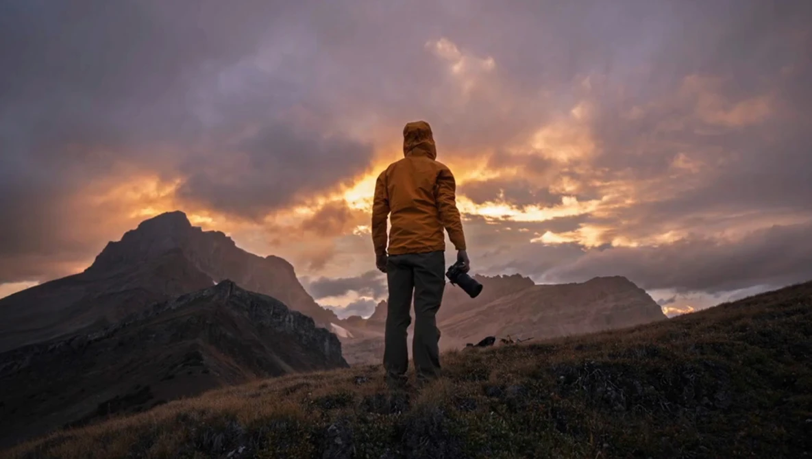 Hiker in yellow jacket standing on mountain terrain at sunset with dramatic clouds.