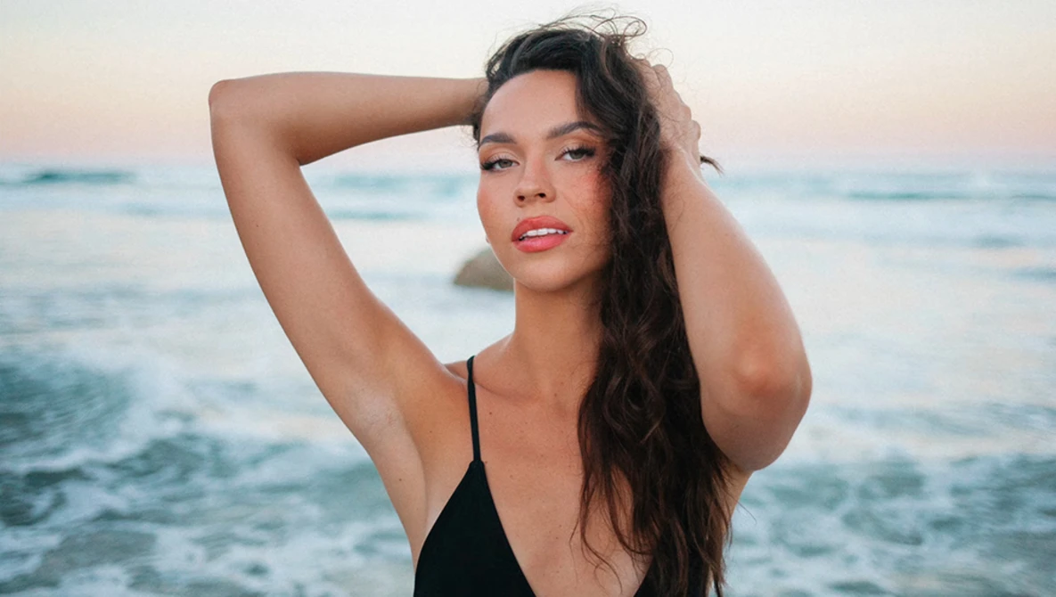 Woman in black swimwear posing at the ocean shore during golden hour sunset.