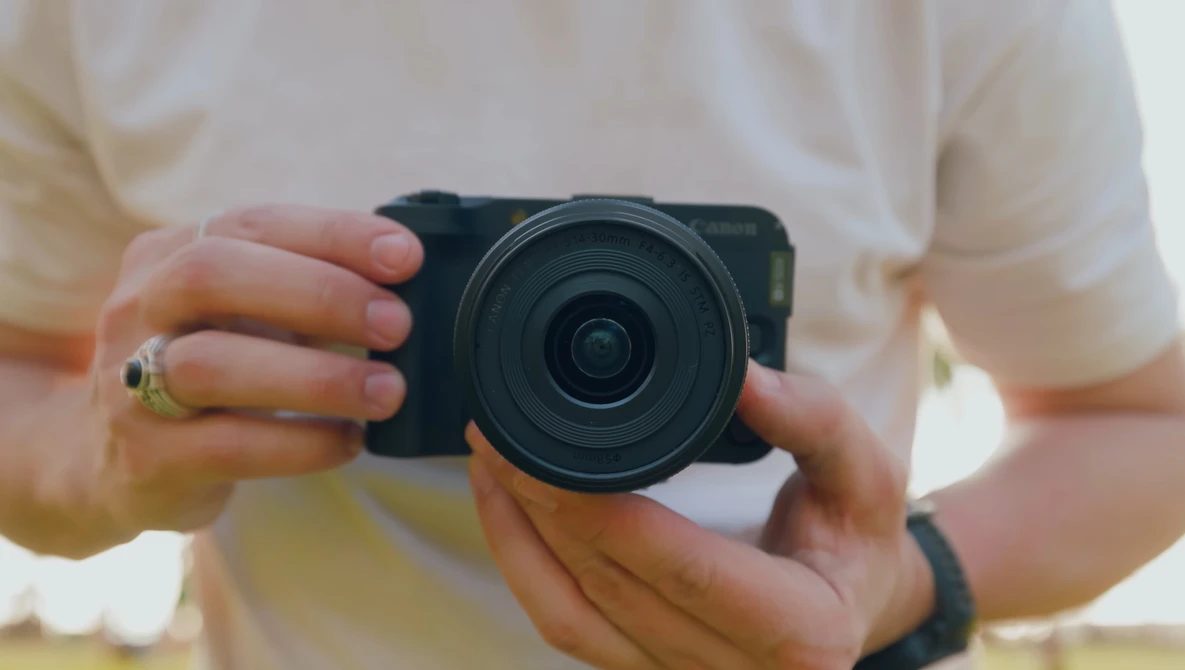 Photographer holding a compact mirrorless camera with a wide-angle lens, wearing a cream-colored shirt and gold ring.