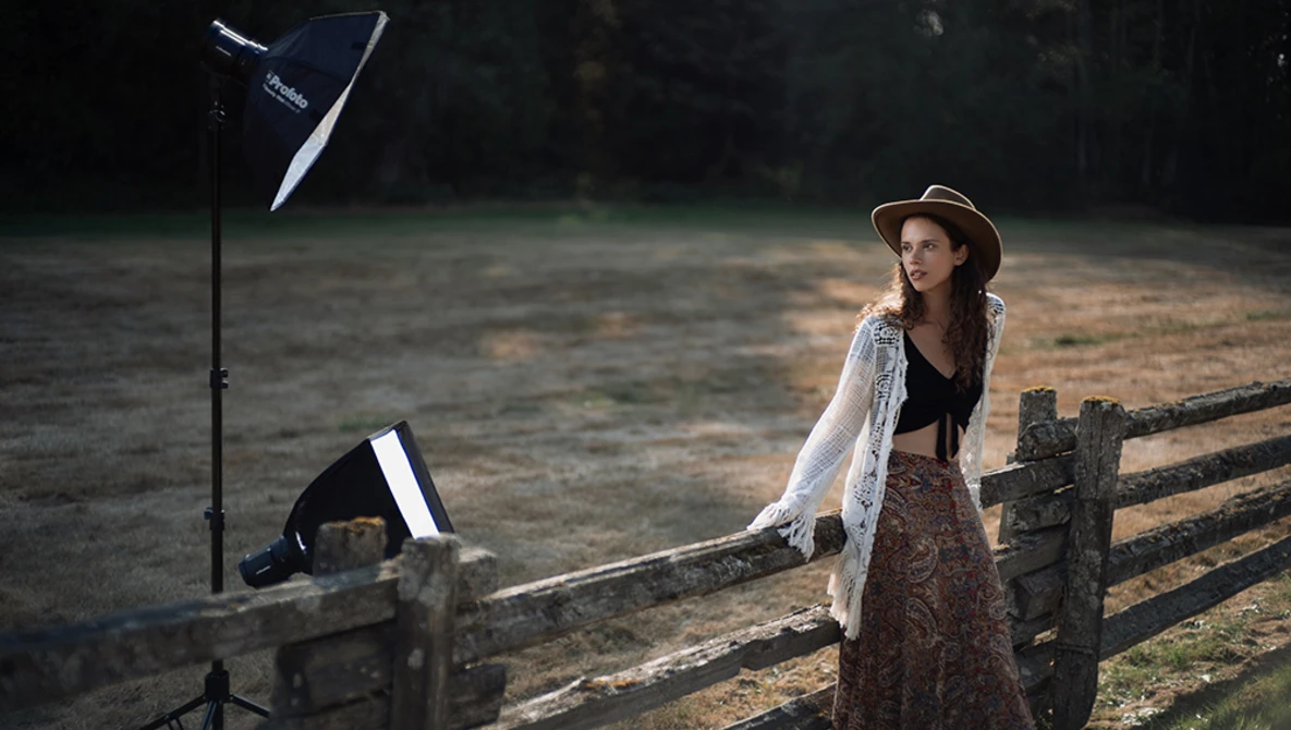 Woman posing by wooden fence with studio lighting setup including reflector and off-camera flash.