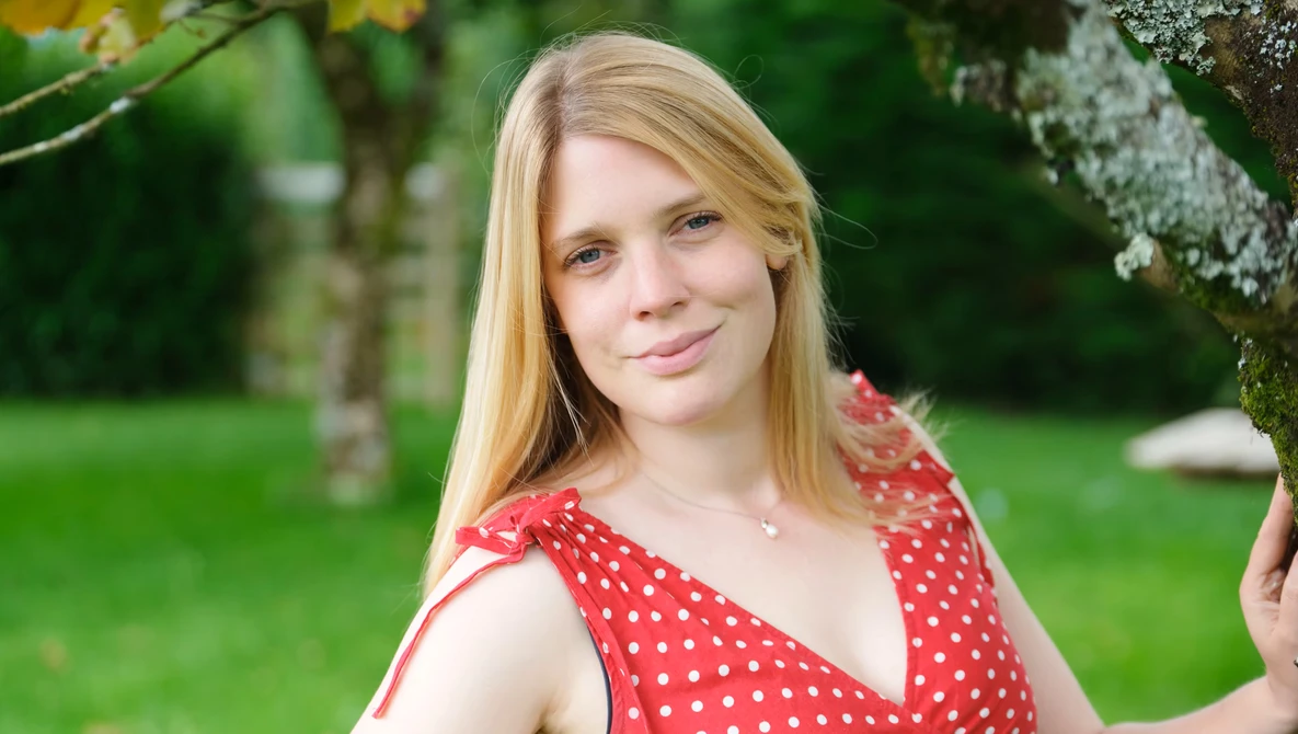 Young woman with blonde hair wearing a red polka dot dress in an outdoor garden setting.