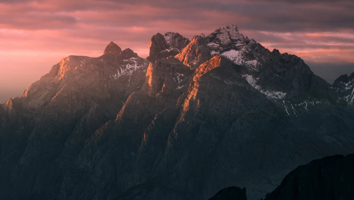 Jagged mountain peaks illuminated by golden alpenglow at sunset against a pink and purple sky.