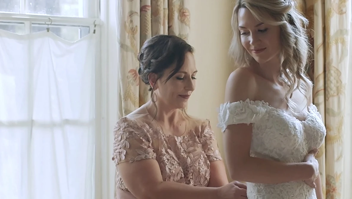 Two women in elegant lace dresses share a moment by a bright window with floral curtains.