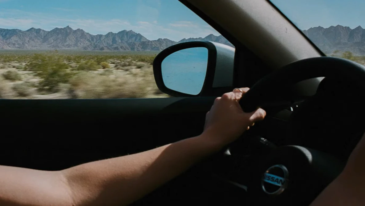 Driver's hand on steering wheel with desert landscape and mountains visible through windshield and side mirror.