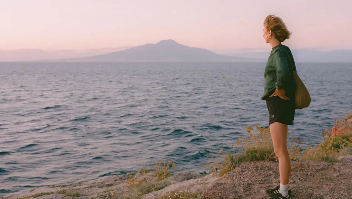 Runner in green jacket standing on coastal cliff at sunset, facing distant snow-capped volcano across water.