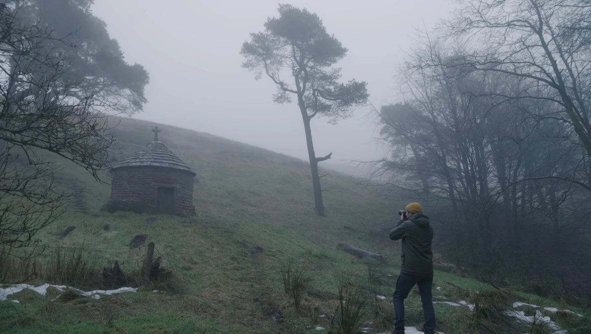 Photographer in yellow beanie shooting in misty hillside landscape with solitary pine tree and stone structure.